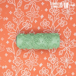 May include: A green textured paint roller with a floral leaf pattern, set against a background of orange paper with a white floral design. The roller has the text "STRUKTUR WALZEN" on the top right.