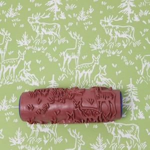 Può includere: Un rullo per pittura strutturato con un motivo di cervi e foresta, nelle tonalità del bordeaux e del blu, poggia su uno sfondo verde con un motivo di cervi e foresta abbinato. Il rullo è progettato per creare motivi decorativi sulle pareti.