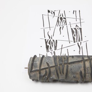 May include: A dark grey paint roller with a textured, geometric design. It sits beneath a black and white abstract pattern on a white backdrop. The roller has a metal rod through the middle.