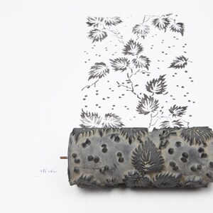Può includere: Un rullo grigio strutturato con un motivo a foglie, utilizzato per stampare un disegno floreale in bianco e nero. Il rullo è posizionato accanto al motivo stampato su carta bianca, mostrando i dettagli del disegno.