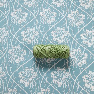 Op de afbeelding: Een groene verfroller met patroon ligt op een blauw bloemenpatroon. De roller heeft een herhalend bloemenontwerp. De achtergrond heeft een herhalend patroon van witte bloemen en wijnstokken op een lichtblauwe achtergrond.