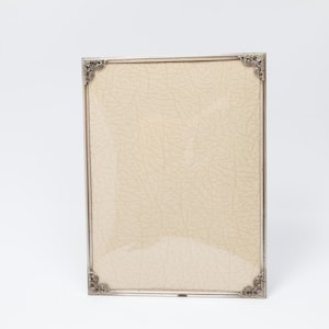 Könnte beinhalten: Ein rechteckiger silberner Bilderrahmen mit verzierten Eckdetails. Der Rahmen enthält einen hellbeige strukturierten Hintergrund. Der Rahmen wird vor einem weißen Hintergrund präsentiert, der sein elegantes Design hervorhebt.