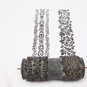 May include: A decorative, dark grey patterned roller with two vertical black and white floral and ornamental designs. The roller has detailed carvings on each end, designed for creating repeating patterns.