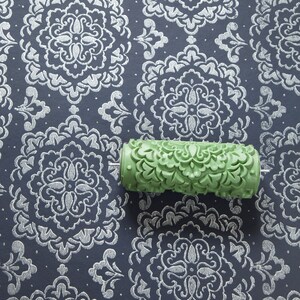 May include: A green patterned paint roller rests on a navy blue surface with a silver damask design. The roller has a detailed floral pattern, suggesting its use for creating textured wall finishes. The background features a repeating, ornate pattern.