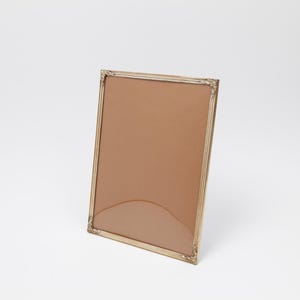 Può includere: Una cornice rettangolare con una cornice in metallo color oro e un retro marrone. La cornice ha accenti decorativi agli angoli. La cornice è appoggiata su uno sfondo bianco.