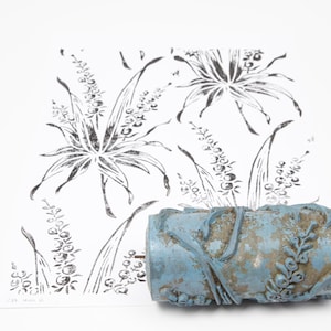 Op de afbeelding: Een verweerde, blauwe verfroller met bloemreliëf ligt voor een zwart-wit bloemenprint. De roller heeft een versleten uiterlijk, wat duidt op ouderdom en gebruik. De print toont gestileerde bloemen en bladeren.