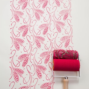 May include: A paint roller with a wooden handle and a red foam roller, next to a wall with a repeating pink leaf pattern. The top roller has a green leaf pattern. The handle has the text "STROKE WALLPAPER".