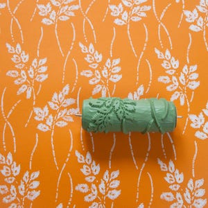 Könnte beinhalten: Eine grüne Farbrolle mit einem Blattdesign. Die Rolle befindet sich auf einem leuchtend orangenen Hintergrund mit einem weißen Blattmuster.