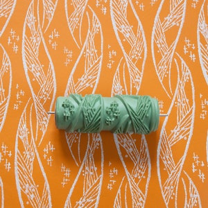 Könnte beinhalten: Eine grüne Farbwalze mit floralem Muster liegt auf einem orangefarbenen Hintergrund mit einem weißen Blättermuster. Die Walze besteht aus einem harten Material und hat eine Metallstange in der Mitte.
