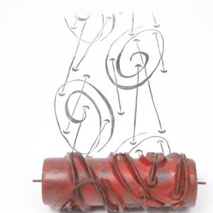 May include: A red patterned paint roller with a metal rod, and a design of grey swirls and arrows above it. The roller is cylindrical and has a textured surface. The background is white.