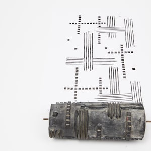 Può includere: Un rullo grigio scuro e strutturato con un motivo geometrico stampato su carta bianca. Il rullo ha una forma cilindrica con vari dettagli scolpiti. Il motivo stampato è composto da linee e croci nere.