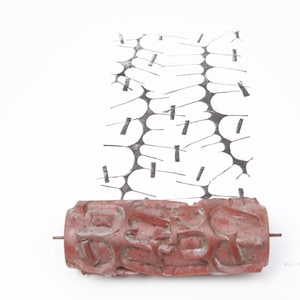 May include: A red, cylindrical object with raised lettering, possibly a printing roller, sits below a black and white abstract design. The roller has a metal rod through its centre. The background is white.