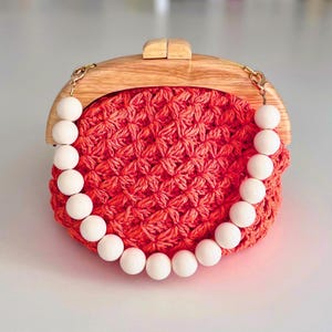 Könnte beinhalten: Eine korallenfarbene gehäkelte Clutch mit Holzrahmen und Verschluss. Die Tasche hat einen weißen Perlenhenkel. Der Holzrahmen ist hellbraun. Die Clutch ist ein stilvolles Accessoire.