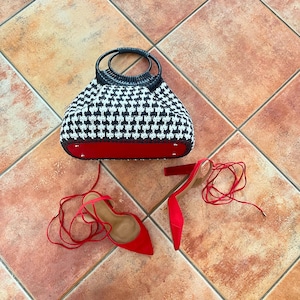 Könnte beinhalten: Eine Handtasche mit schwarz-weißem Hahnentrittmuster mit rotem Boden und schwarzen Griffen. Daneben ein Paar rote Wildleder-High Heels mit Schnürung. Die Artikel werden auf einer gefliesten Oberfläche präsentiert.