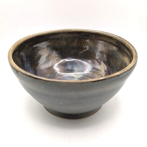 Peut inclure: Un bol en céramique fait à la main avec un intérieur sombre et brillant et un bord plus clair. Le bol a un extérieur lisse et gris avec de subtiles variations de couleur. L'intérieur reflète la lumière, présentant un mélange de bleus foncés, de bruns et d'ors.