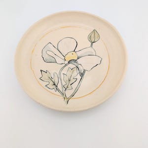 May include: A white ceramic plate with a hand-painted design of a white flower with a yellow center and green leaves. The plate has a light brown rim.