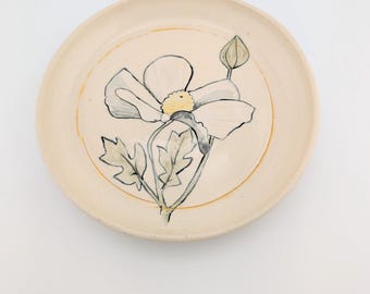 May include: A white ceramic plate with a hand-painted design of a white flower with a yellow center and green leaves. The plate has a light brown rim.