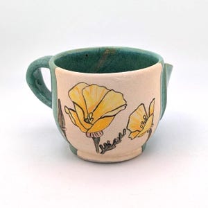 Może przedstawiać: Ceramiczny dzbanuszek do śmietanki z turkusowym uchwytem i obramowaniem. Korpus jest jasnoróżowy z ilustracjami żółtych kwiatów maku. Kwiaty mają czarne kontury i zielone łodygi. Dzbanuszek ma mały dzióbek.