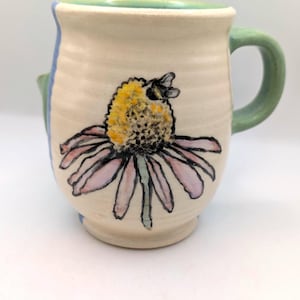 Puede incluir: Una taza de cerámica hecha a mano con asa y borde verdes, y un acento azul. La taza presenta un diseño pintado a mano de una abeja sobre una equinácea con pétalos rosas y centro amarillo. La taza es de color blanquecino.