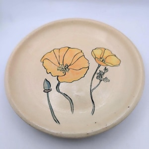 Könnte beinhalten: Eine Keramikschale mit handgemaltem Design von gelben und orangefarbenen Mohnblumen auf cremefarbenem Hintergrund.