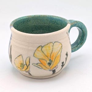 Pode incluir: Caneca de cerâmica artesanal com exterior creme e interior e alça turquesa. Apresenta papoulas amarelas pintadas à mão com caules e folhas pretas. Uma peça artística única para desfrutar de bebidas.