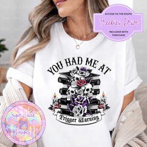 May include: White t-shirt with a black and white graphic design featuring a stack of books with skulls and roses. The text "You Had Me At Trigger Warning" is printed on the shirt.