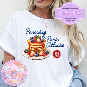 May include: White t-shirt with a graphic of a stack of pancakes with strawberries and blueberries, topped with syrup. The text "Pancakes & Panic Attacks" is written in blue. A red circle with the text "Buy 1 Get 1 Free" is in the lower right corner.