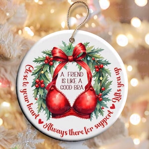 May include: A white ceramic ornament with a red and green wreath and a red bra. The ornament has the text "Close to your heart Always there for support Lifts you up" and "A friend is like a good bra".