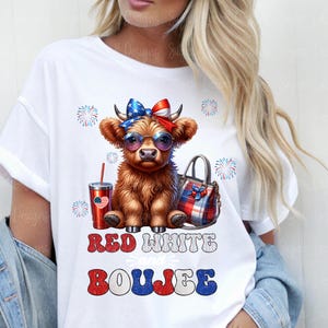 May include: A white t-shirt featuring a graphic of a brown Highland cow wearing a red, white, and blue bow and sunglasses. Beside the cow is a red tumbler and a plaid handbag. The text reads RED WHITE and BOUJEE with firework accents.