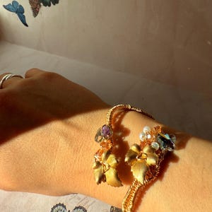 May include: Gold-toned cuff bracelet featuring floral and leaf accents. The bracelet is adorned with faceted crystals and small pearls. The design incorporates a twisted wire structure, adding texture and visual interest to the jewelry.