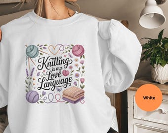 May include: White sweatshirt featuring the phrase "Knitting is my Love Language" in a decorative font, surrounded by yarn balls, flowers, and a stack of folded blankets. The design includes shades of purple, pink, and green.