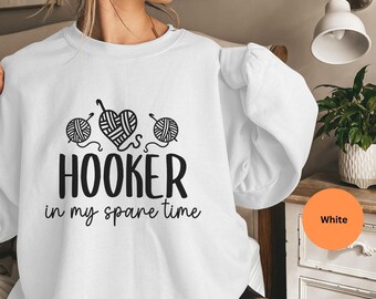 May include: White sweatshirt with the text "HOOKER in my spare time" in black, along with illustrations of yarn balls and a crochet hook forming a heart. The sweatshirt has long sleeves.