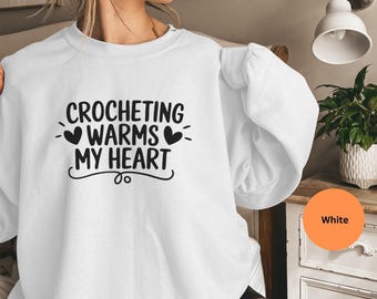 May include: A white sweatshirt with the text "CROCHETING WARMS MY HEART" in black. The design includes heart shapes. The sweatshirt is a casual garment, suitable for crafters and those who enjoy crochet.