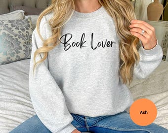 May include: Light gray sweatshirt with the text "Book Lover" in black script. The sweatshirt is a crew neck style and is made of a soft material. The color is ash.