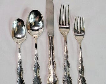 May include: A set of five silver-colored flatware pieces, including a spoon, a large spoon, a knife, a fork, and a smaller fork. Each piece has a decorative handle with a floral design.