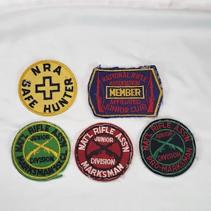 May include: A collection of vintage patches with text and graphics. One patch reads "NRA SAFE HUNTER" with a cross. Other patches say "NATIONAL RIFLE ASSOCIATION MEMBER" and "NAT'L RIFLE ASS'N DIVISION MARKSMAN".