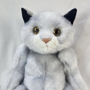 Peut inclure: Une peluche en forme de chat avec une fourrure douce gris clair, des oreilles pointues aux extrémités noires et de grands yeux ambrés réfléchissants. Le visage présente un museau blanc, un petit nez rose et de fines moustaches blanches.