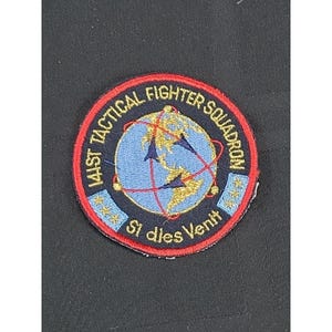 May include: Embroidered patch with a red border, featuring the text "141ST TACTICAL FIGHTER SQUADRON" in white. The center shows a blue globe with gold continents and red lines. The text "Si dies Venit" is at the bottom.