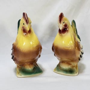 May include: A pair of ceramic chicken figurines featuring a yellow head and neck, a brown chest and body, and a green base. Each chicken has a dark red comb and wattle, with dark green tail feathers visible on the back. The figures are standing upright.