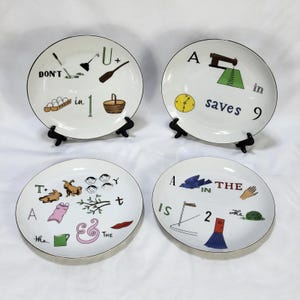 Può includere: Un set di quattro piatti in ceramica bianca con illustrazioni grafiche colorate e testo. I disegni includono giochi di parole visivi e frasi come 'DON'T U + in 1', 'A in saves 9', 'A T+ Y' e 'A IN THE IS 2 THE' con varie icone come un cestino, un orologio e una macchina da cucire.