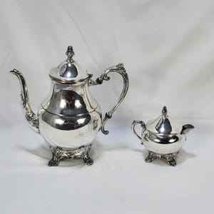 Op de afbeelding: Een gepolijste zilveren theepot en een bijpassende suikerpot met deksels. Beide stukken zijn voorzien van sierlijke, gebogen handvatten, decoratieve knoppen op de deksels en ingewikkelde, krullende voetjes. De theepot heeft een lange tuit.