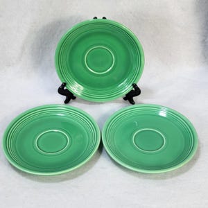 Pode incluir: Três pires verdes de cerâmica. Dois estão planos e um está elevado em um suporte preto. Os pires têm uma reentrância circular no centro e anéis concêntricos na borda. A cor é verde brilhante e sólida.