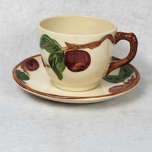 May include: Cream-colored teacup and saucer set with a hand-painted apple design. The cup has a brown handle and the saucer has a matching brown rim. The apple design features red apples, green leaves, and brown branches.