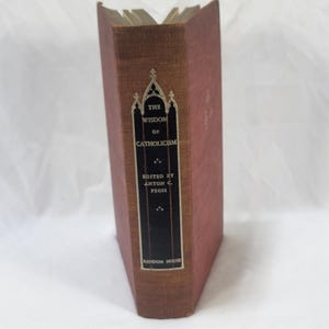 May include: A hardcover book titled The Wisdom of Catholicism edited by Anton C. Pegis and published by Random House. The book features a textured, reddish-brown fabric cover and a black spine label with white text and a gothic arch design.
