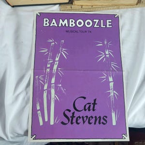 May include: Purple vintage poster for the "Bamboozle Musical Tour '74" featuring Cat Stevens. The poster has a white border and includes bamboo illustrations. The text is in white and black, with the artist's name in a stylized font.