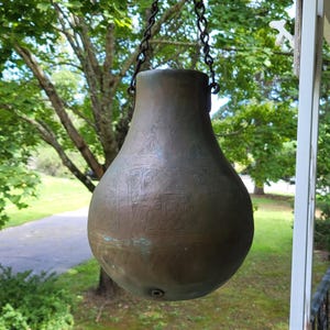 May include: A pear-shaped, dark bronze-toned metal vessel with a wide, rounded base and a narrow neck. The surface features intricate, etched geometric and floral patterns. It is suspended by a dark metal chain attached to two side loops.