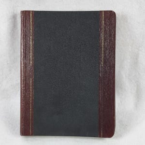 Op de afbeelding: Een gesloten, rechthoekig boek met een donkergrijze kaft en bordeauxrode rug en randen. De rug en randen hebben een gouden verticale streep. Het boek ligt op een wit oppervlak.