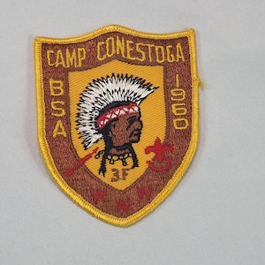 Puede incluir: Un parche vintage del Campamento Conestoga BSA. El parche en forma de escudo tiene un borde dorado y un fondo marrón. Presenta un jefe nativo americano con un tocado de plumas, el logotipo de BSA y el texto "Camp Conestoga 1960".