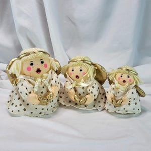 May include: Three plush angel dolls of varying sizes, each featuring cream-colored fabric with a brown star pattern, gold metallic halos, and gold wings. The dolls have yarn hair, painted facial features with pink cheeks, and hold gold hearts.