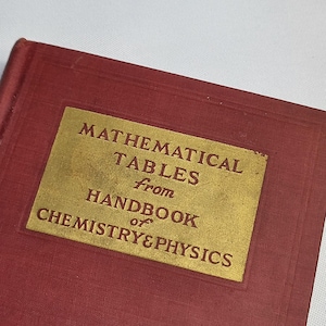 Puede incluir: Un libro antiguo con una cubierta granate y una etiqueta dorada. La etiqueta dice "MATHEMATICAL TABLES from HANDBOOK of CHEMISTRY & PHYSICS". El libro está ligeramente desgastado, lo que sugiere edad y uso.